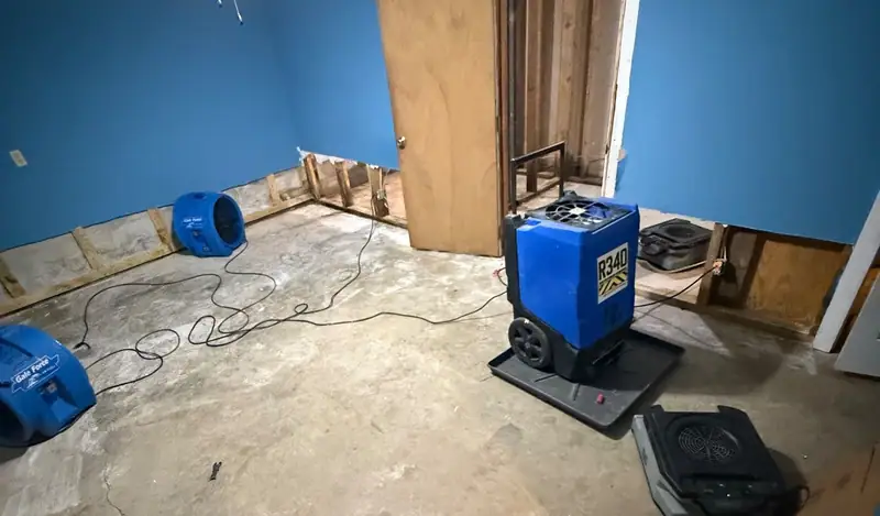 Blue bedroom mid-dry-out: drywall cut at the floor line, a dehumidifier and air movers running